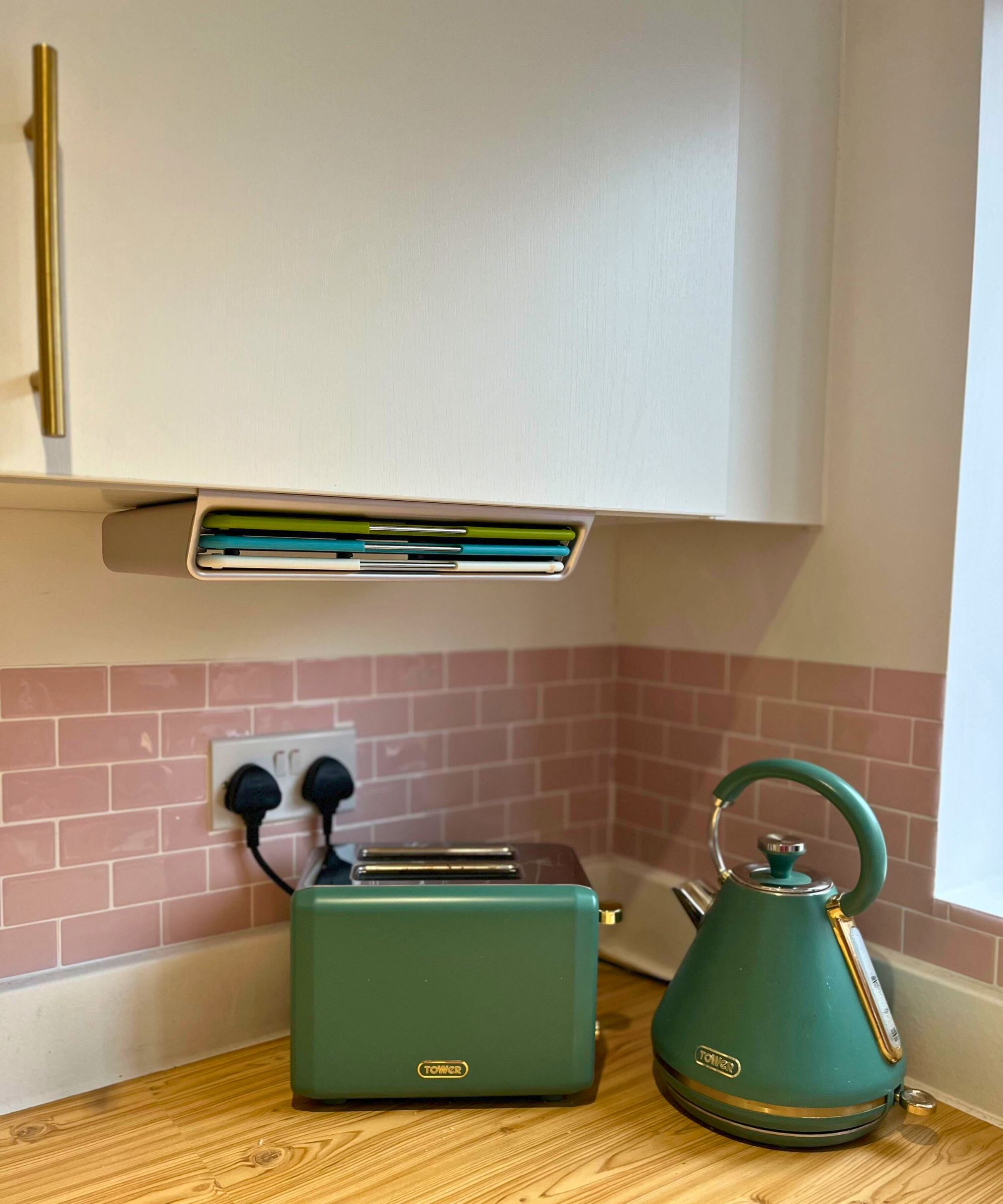Collect photo of Joseph Joseph under-shelf caddy attached under a wall cabinet, with and trio of cutting boards visible in white, blue and green. There is a green toaster and kettle on wooden kitchencounters underneath.