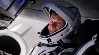 andreas mogensen in a spacesuit sitting in a spacecraft, looking at camera and smiling. screens are visible to the upper left of image
