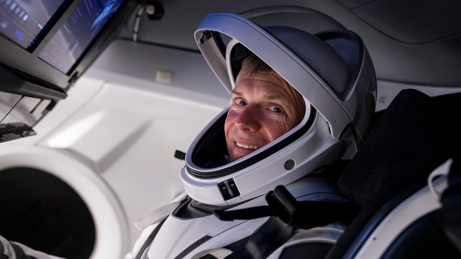 Andreas Mogensen ve skafandru sedí v kosmické lodi, dívá se do kamery a usmívá se.  Obrazovky jsou viditelné v levé horní části obrázku