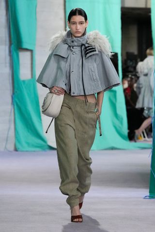 A woman wears a grey cape jacket and green pants on Burberry's Spring 2025 runway.