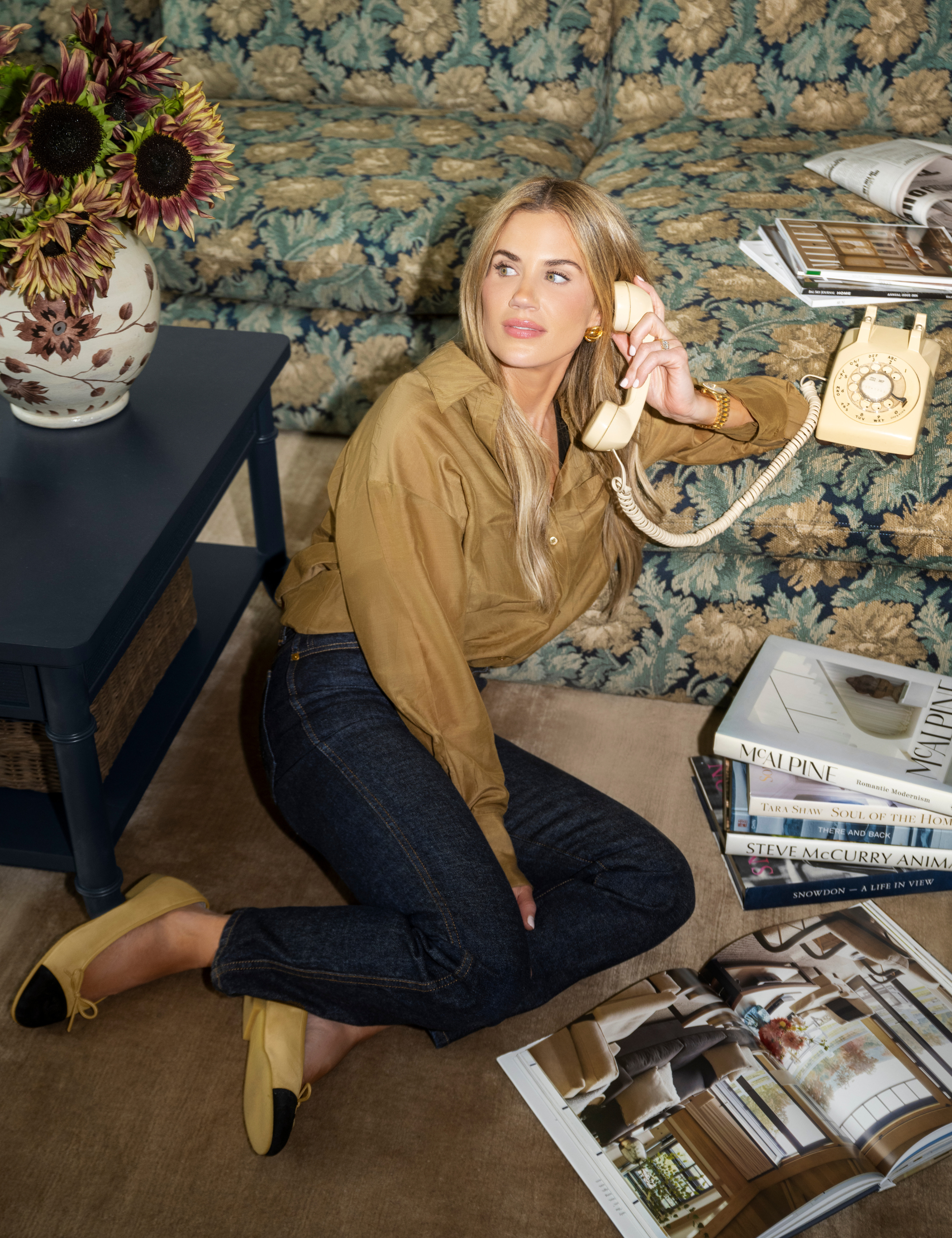 Shea McGee poses holding a vintage dial phone on a carpeted floor alongside a jacquard floral sectional