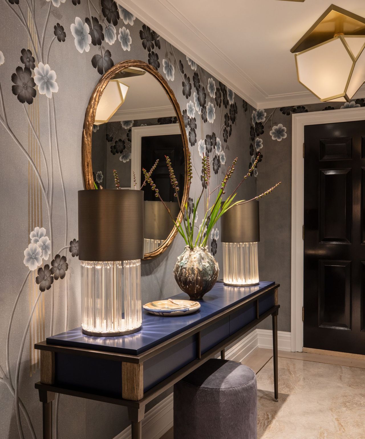 An entryway with a dark blue console table, two lamps, and a round mirror against blue wallpaper