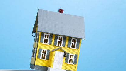 Miniature model of yellow single family house about to fall off edge of white shelf, blue background