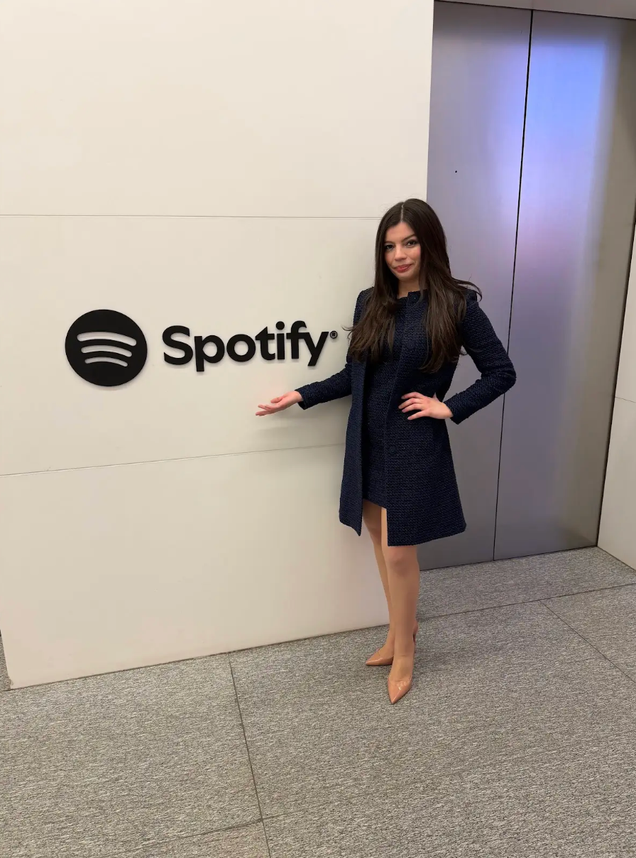 Author Alex Aster poses outside the Spotify offices wearing a navy dress and matching jacket with nude heels.