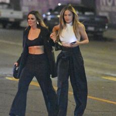 Margot Robbie walks across the road wearing black high-waisted trousers with black platform leather heels and a white sleeveless top.