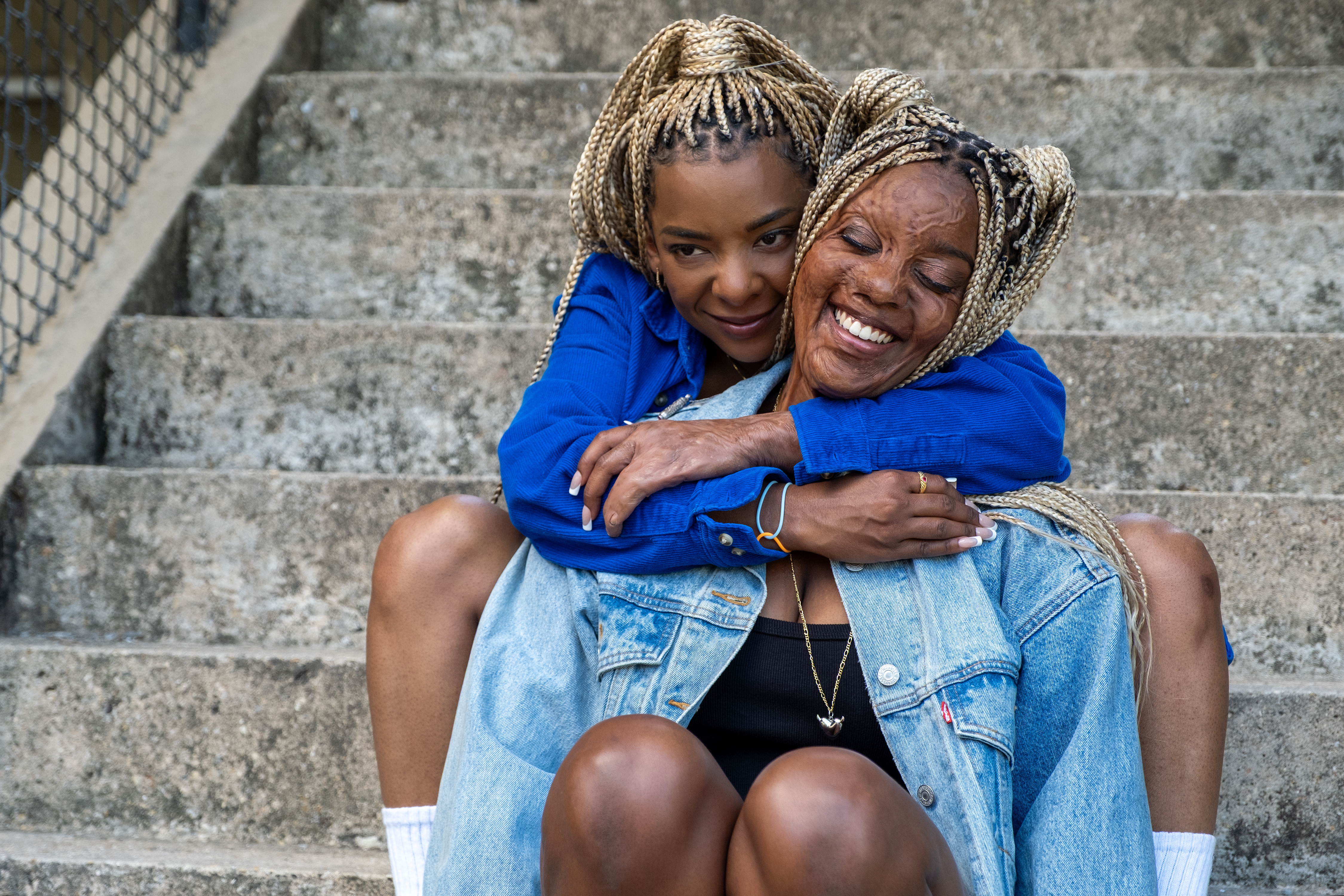 A woman with blonde box braids (Kara Young as Racine) back-hugs a woman with facial scars (Mallori Johnson as Anaia) as they both sit on outdoor steps, in a scene from 'Is God Is.'