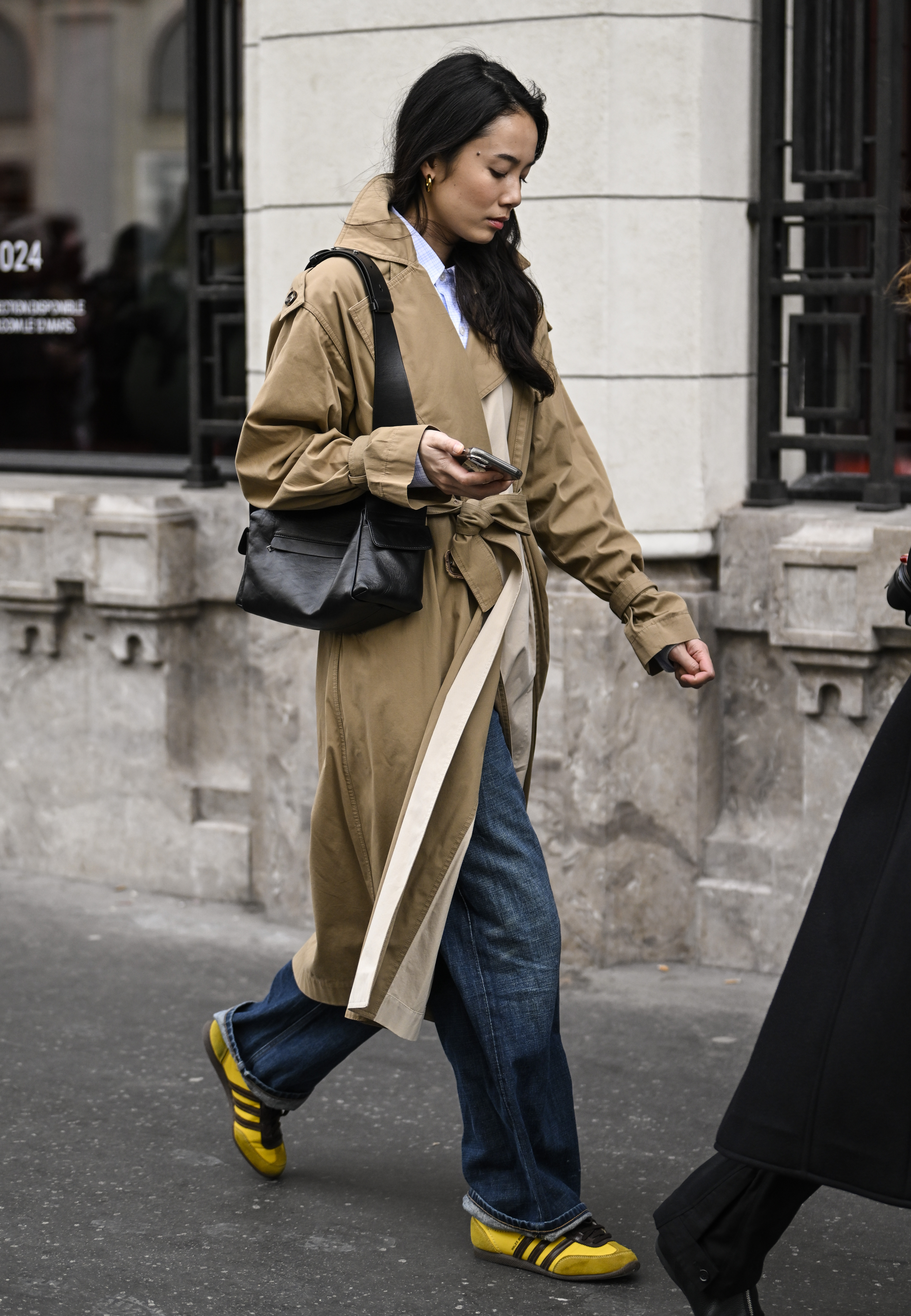 PARIS, FRANCE - FEBRUARY 28: Celine Duong is seen wearing a tan and cream trench coat, blue jeans and yellow and black sneakers with a black bag outside the Dries Van Noten show during the Womenswear Fall/Winter 2024/2025 as part of Paris Fashion Week on February 28, 2024 in Paris, France. (Photo by Daniel Zuchnik/Getty Images)