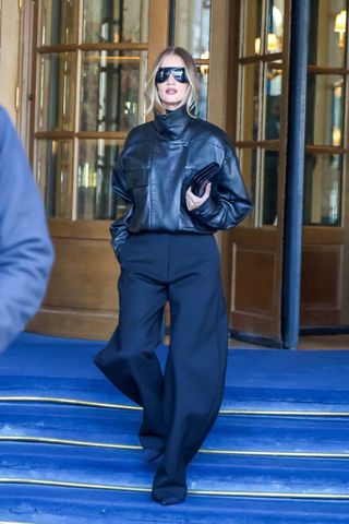 Rosie Huntington Whiteley wearing a leather funnel-neck jacket with barrel leg trousers, heels, a clutch, and oversized sunglasses outside of the Ritz Hotel in Paris during Paris Fashion Week.