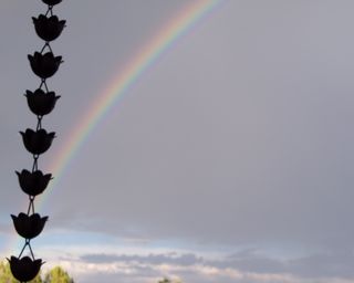 A rain-chain that directs water off of a roof in silhouette and the rainbow against a cloudy sky after a rain storm