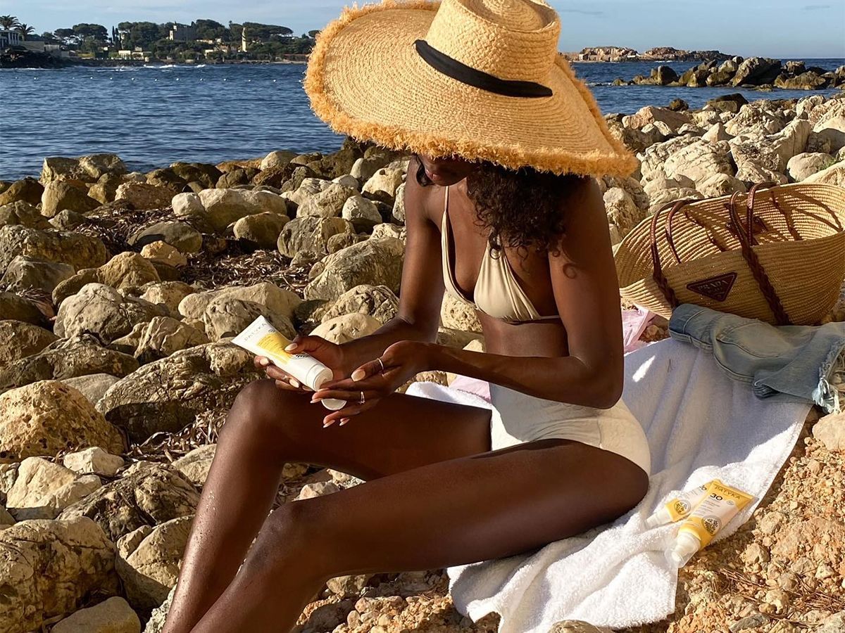 A woman sitting at the beach wearing a white bikini set and a large straw hat.