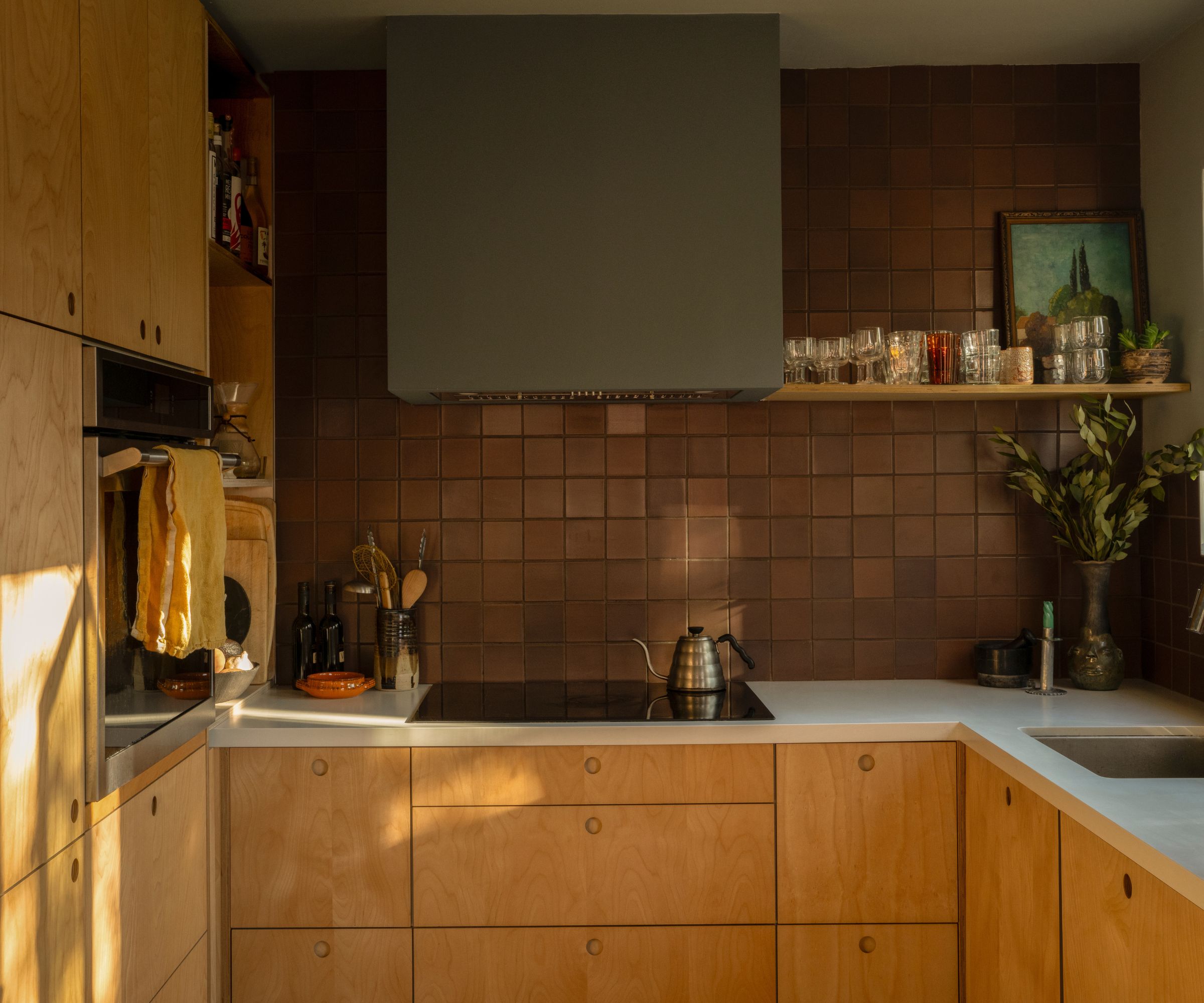 Close-up of a kitchen corner with light wood grain cabinets, a dark brown square tile backsplash, and a large matte grey range hood.