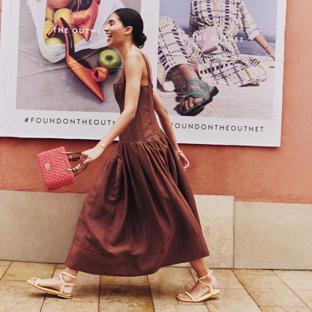Young brunette woman wearing brown dress, white sandals, pink handbag from The Outnet, walking past The Outnet posters.
