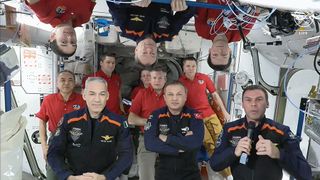 11 astronauts, 7 in red shirts and 4 in blue and orange flight suits on the space station. some are upside down