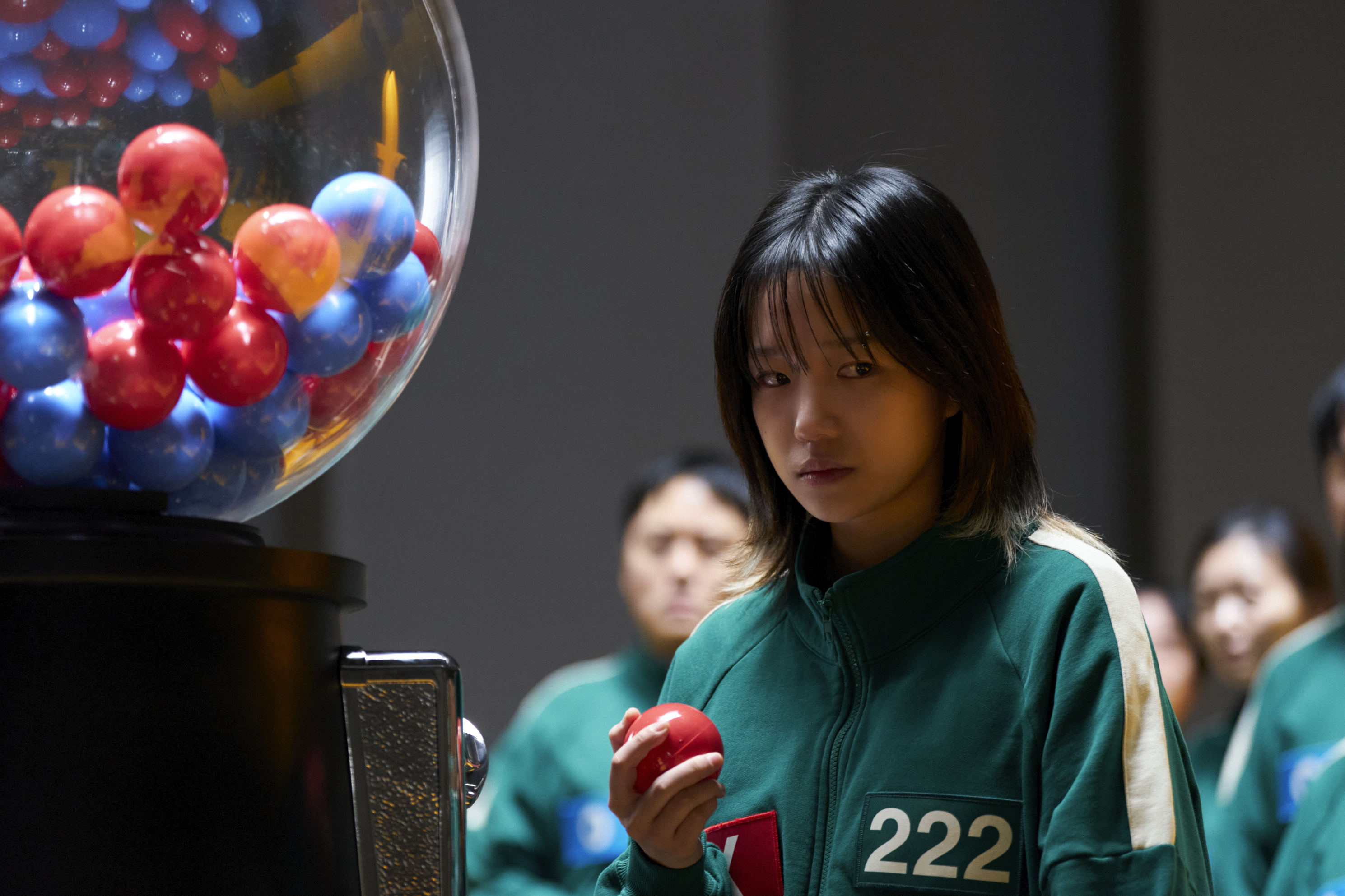 A woman (Jo Yu-ri as Jun-hee, Player 222) holds a ball from a large gumball machine, in 'Squid Game' season 3.