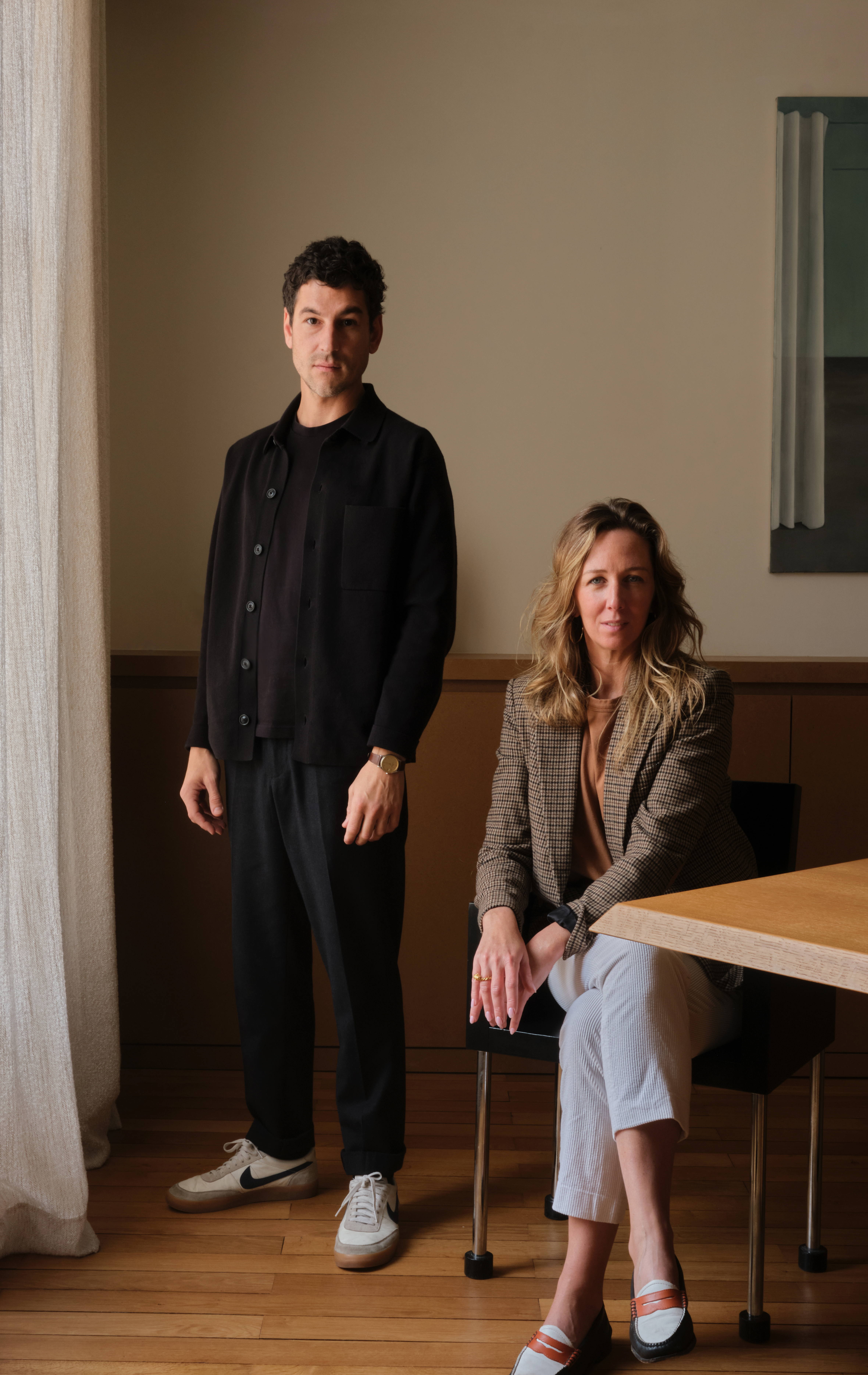 A portrait of a designer duo composed of a man dressed in a total black, casual-chic outfit, and a woman, sitting next to him, in a checkered blazer in brown and white trousers.