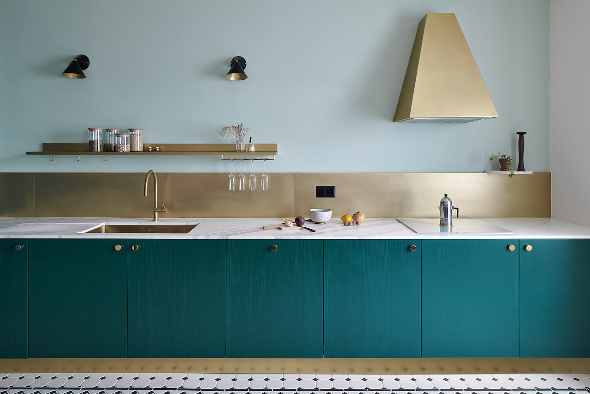 a modern kitchen with teal kitchen cabinets, a bronzed backsplash and kickboard, sculptural rangehood and floating shelf holding cups and plates. there is a sink with silver tap, and two wall sconces overhead
