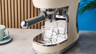 a cream-colored retro espresso machine by italian brand smeg is photographed against a blue background with an empty shot glass awaiting espresso