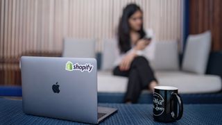 Photograph of laptop with Shopify sticker, on table beside coffee mug, with woman on her phone out of focus in background