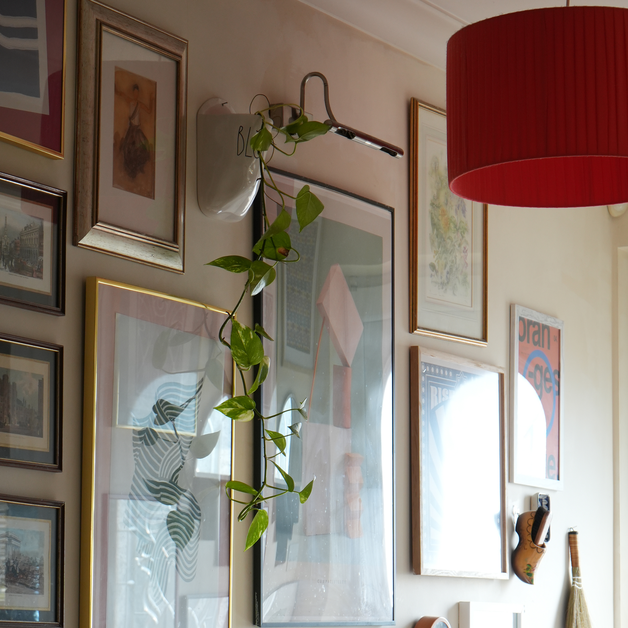 gallery wall of artwork framed in a variety of frames, on a pale wall with a red pendant light next to it