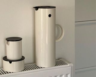 Minimal off-white tall slim jug with handle and black lid resting on kitchen radiator alongside matching mug with lid