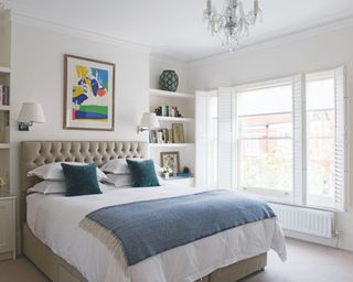 A large double bedroom with a buttoned headboard bed with white bedding, a blue blanket and blue cushons, white walls with colorful framed artwork above the bed, shelves with miscellaneous items such as books, framed pictures and vases, and large windows with shutters. A glass chandelier hangs above