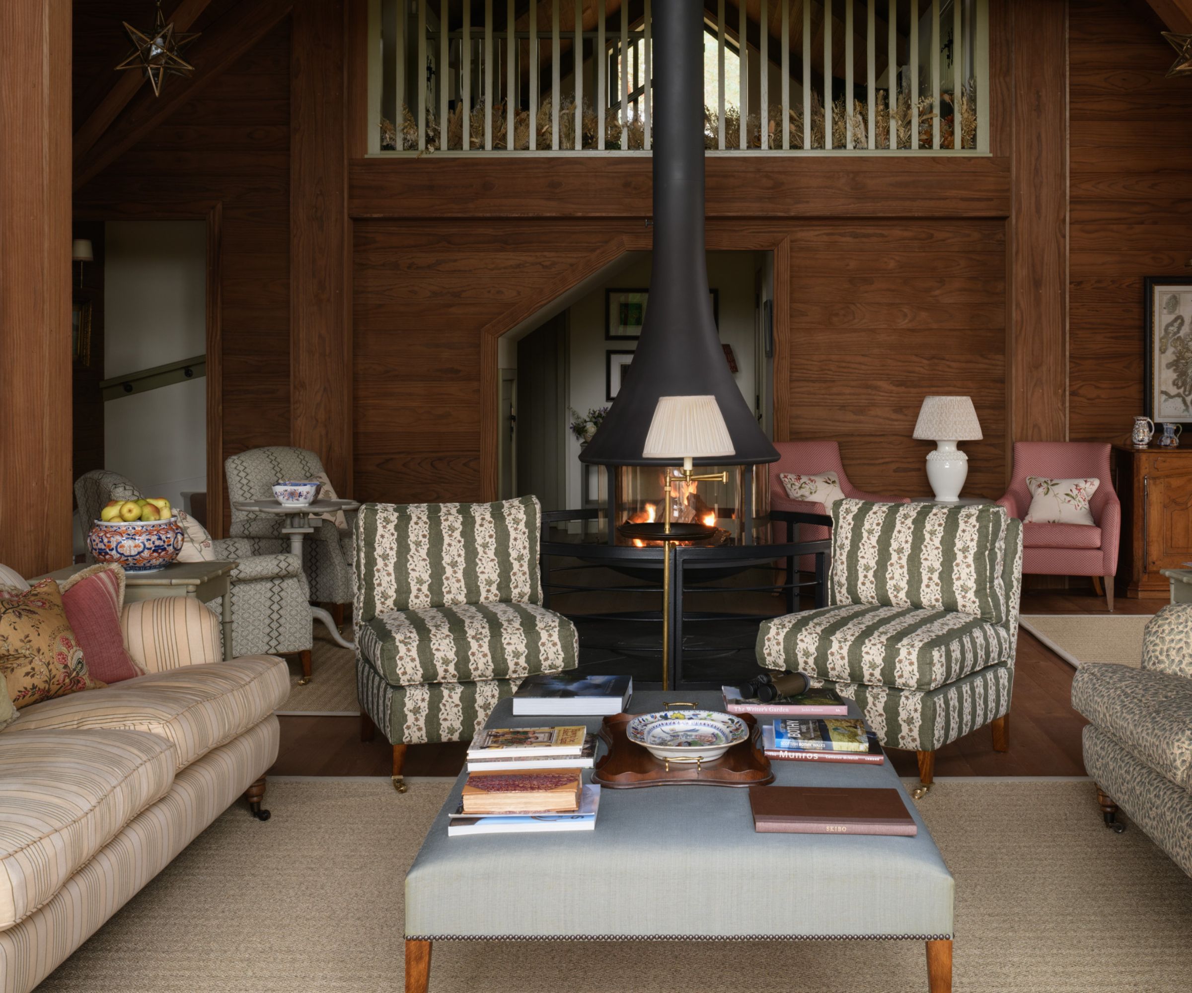 A warm, character-filled living room in a woodland lodge, complete with eclectic upholstery, a central fireplace and a gallery mezzanine above. Timber walls and soft neutral tones give the space a relaxed, heritage feel.