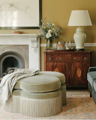 A sage green fringed ottoman in a traditional living room with a dresser and marble surround fireplace