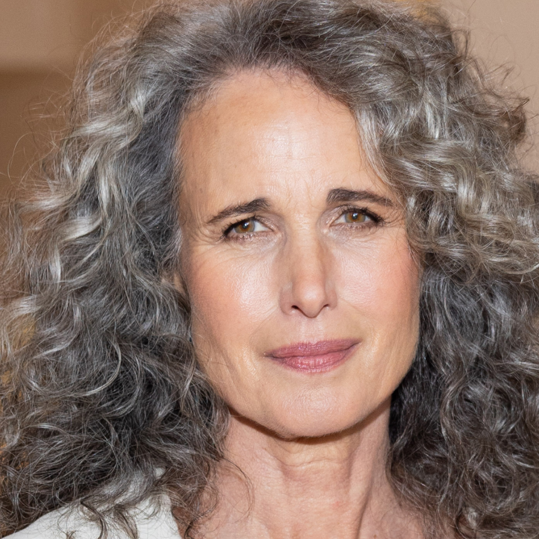 Andie MacDowell with curly hair and skin-like makeup on, posing for the camera. 