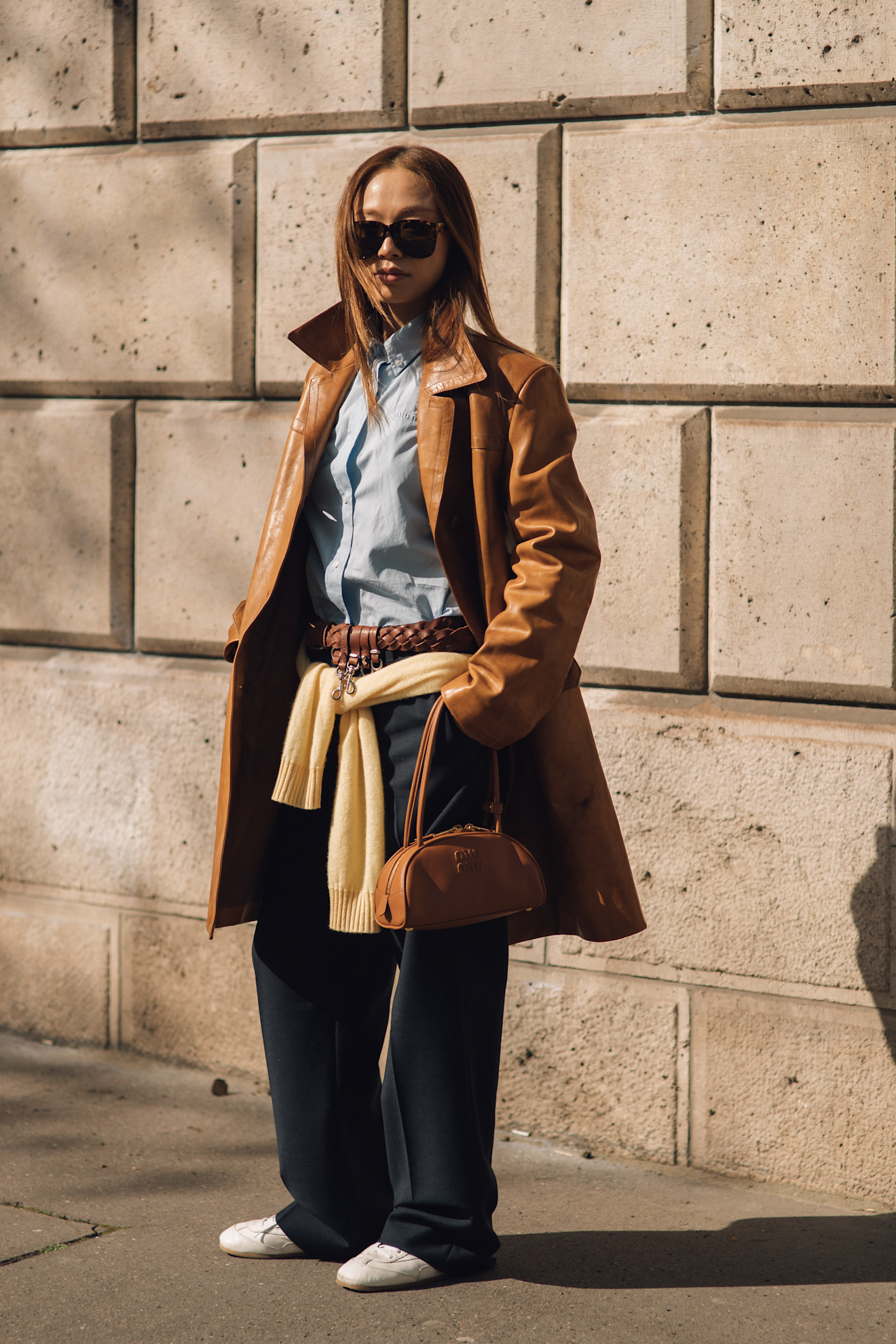 fashion week attendee wearing sunglasses, tan leather jacket, blue shirt, trousers, and white sneakers
