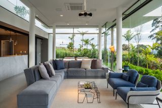 Interior view of the lounge room. Thin white columns support the ceiling. A large grey sofa and navy blue and gray armchairs are turned toward all glass walls, which provide a view of the beautiful nature surrounding the house.