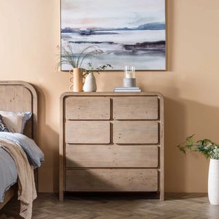 Contemporary wooden chest of drawers next to a bed in a peach bedroom with wood floors