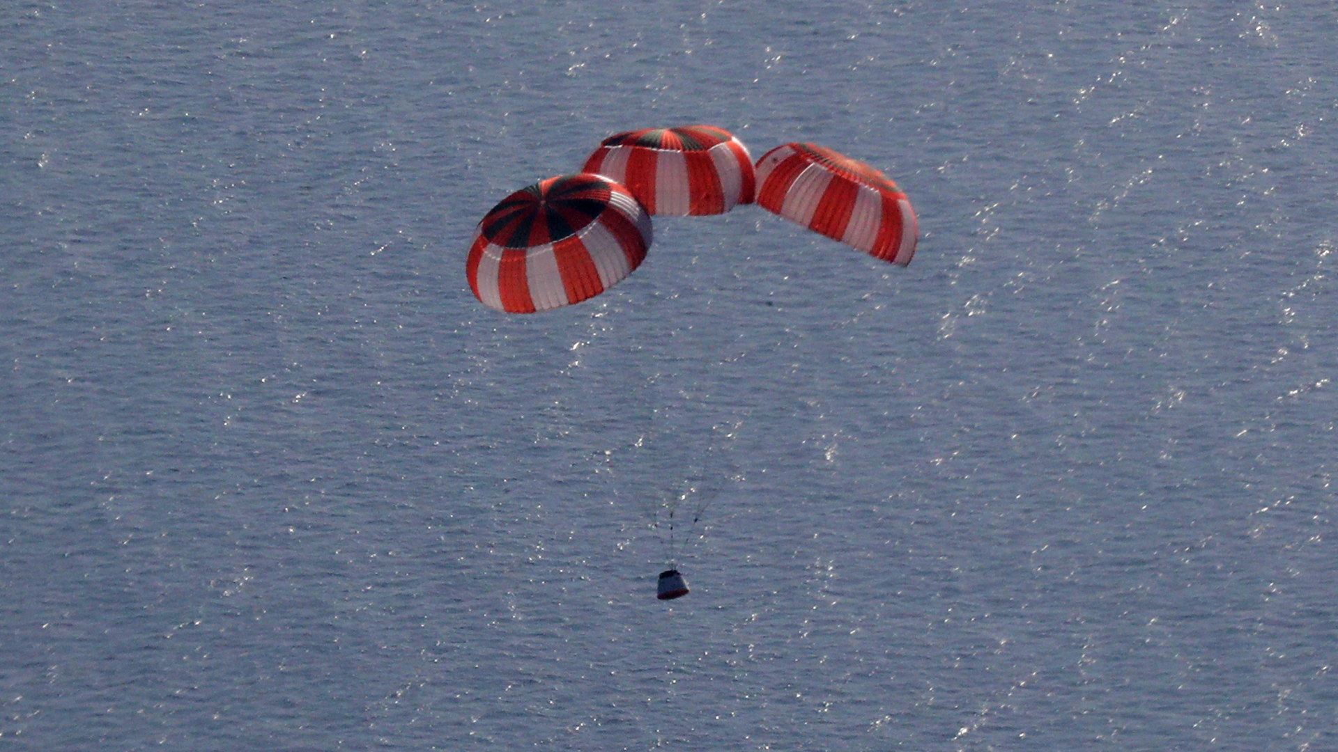Three red and white parachutes carry a large metal crew capsule to a safe landing in the ocean
