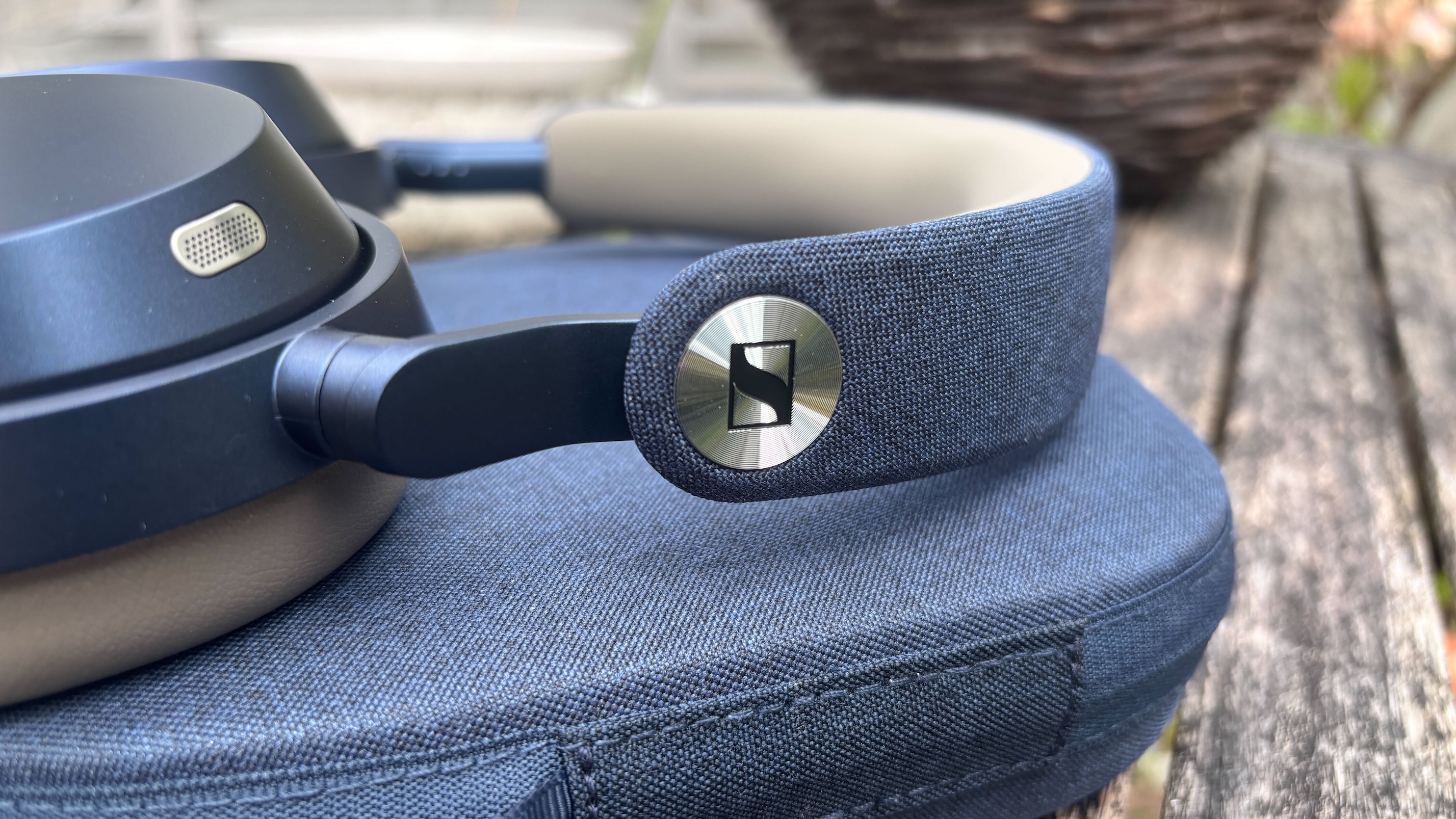 A close-up of the Sennheiser logo on the headband of the Sennheiser Momentum 5 Wireless headphones, which are lying on their case on a table outside.