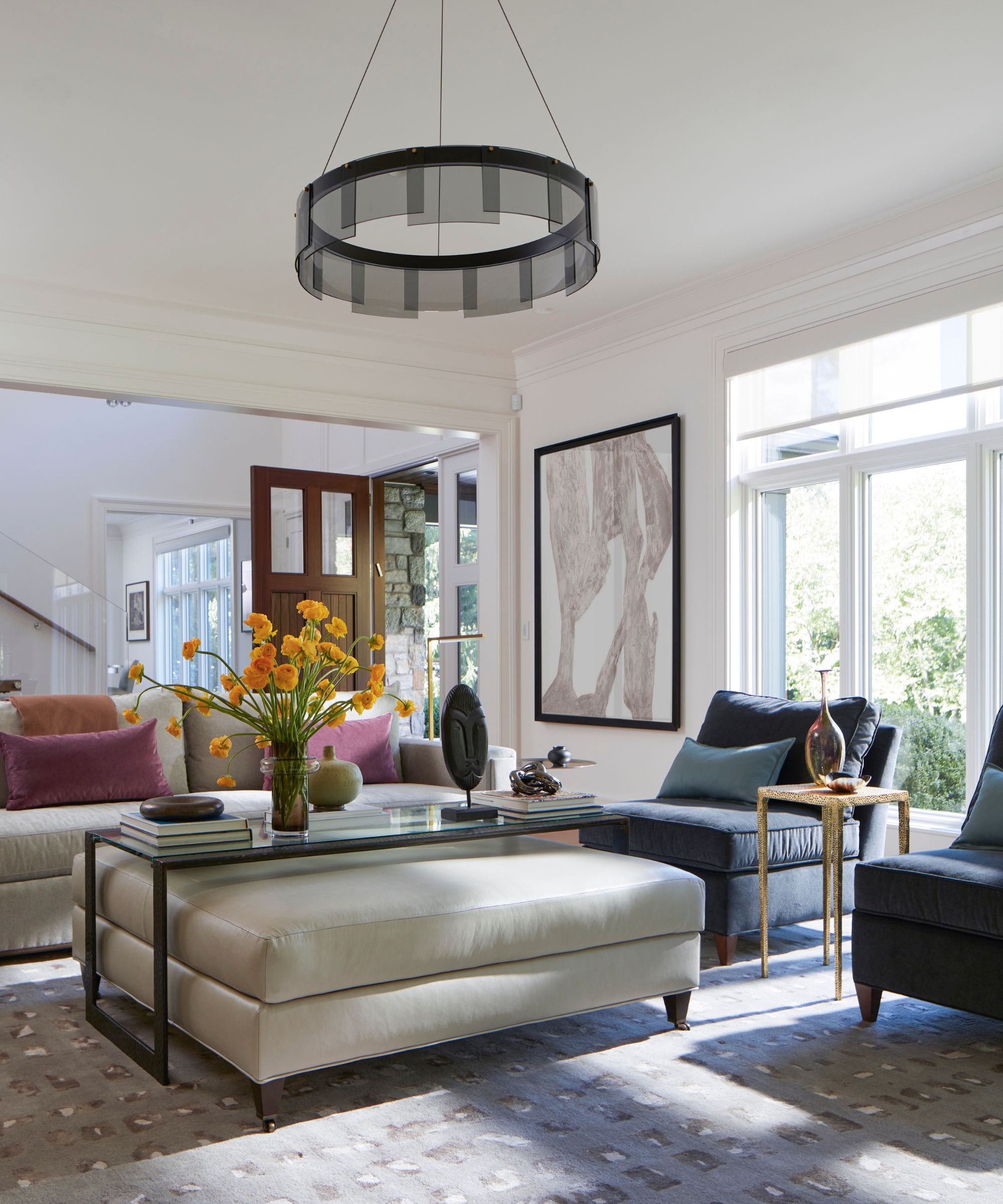 Living room with large modern pendant light, white upholstered ottoman, navy armchairs and cream sofa with purple pillows