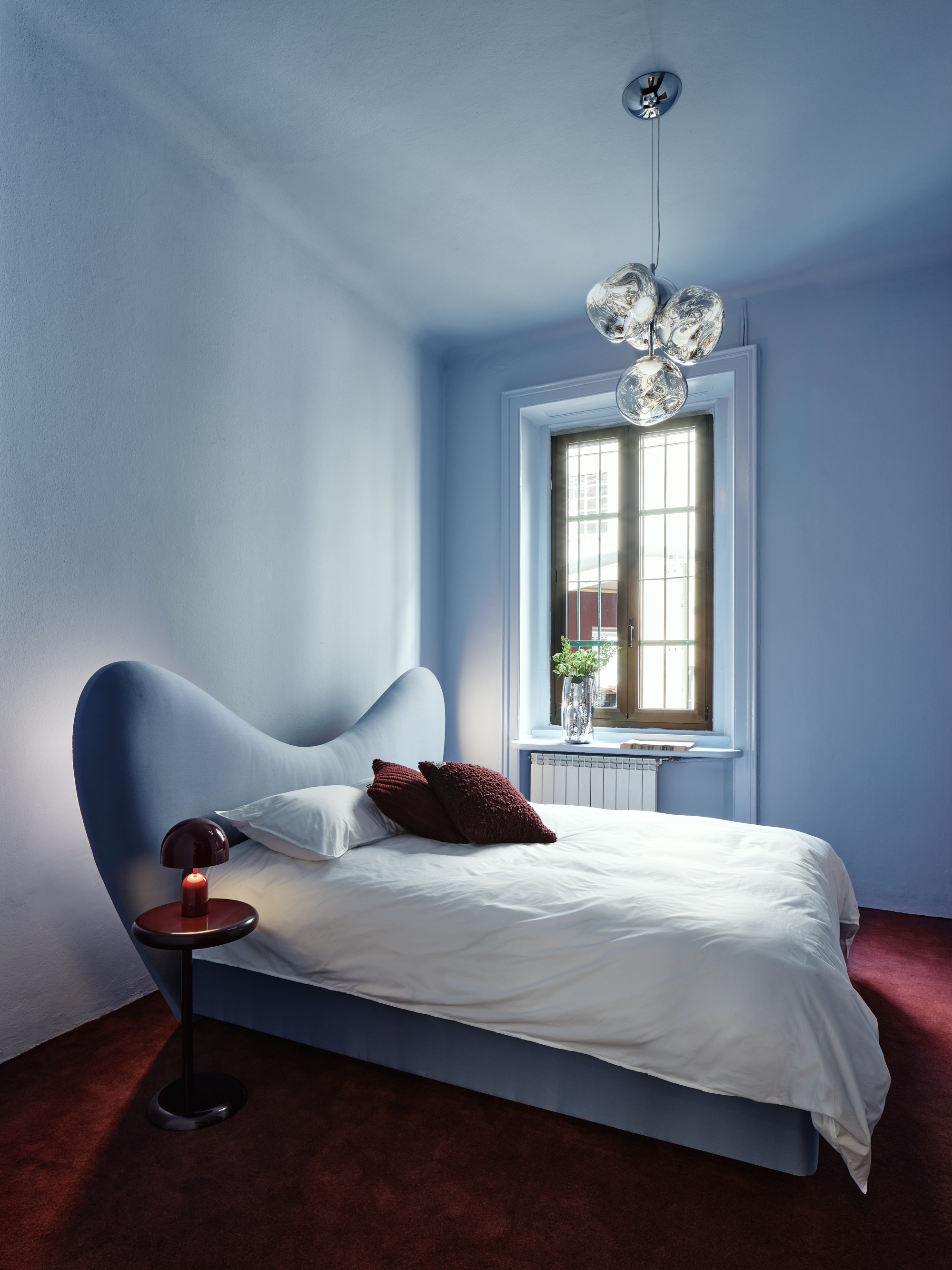 A light blue bedroom with oxblood red carpet with a boldly shaped, curved bed