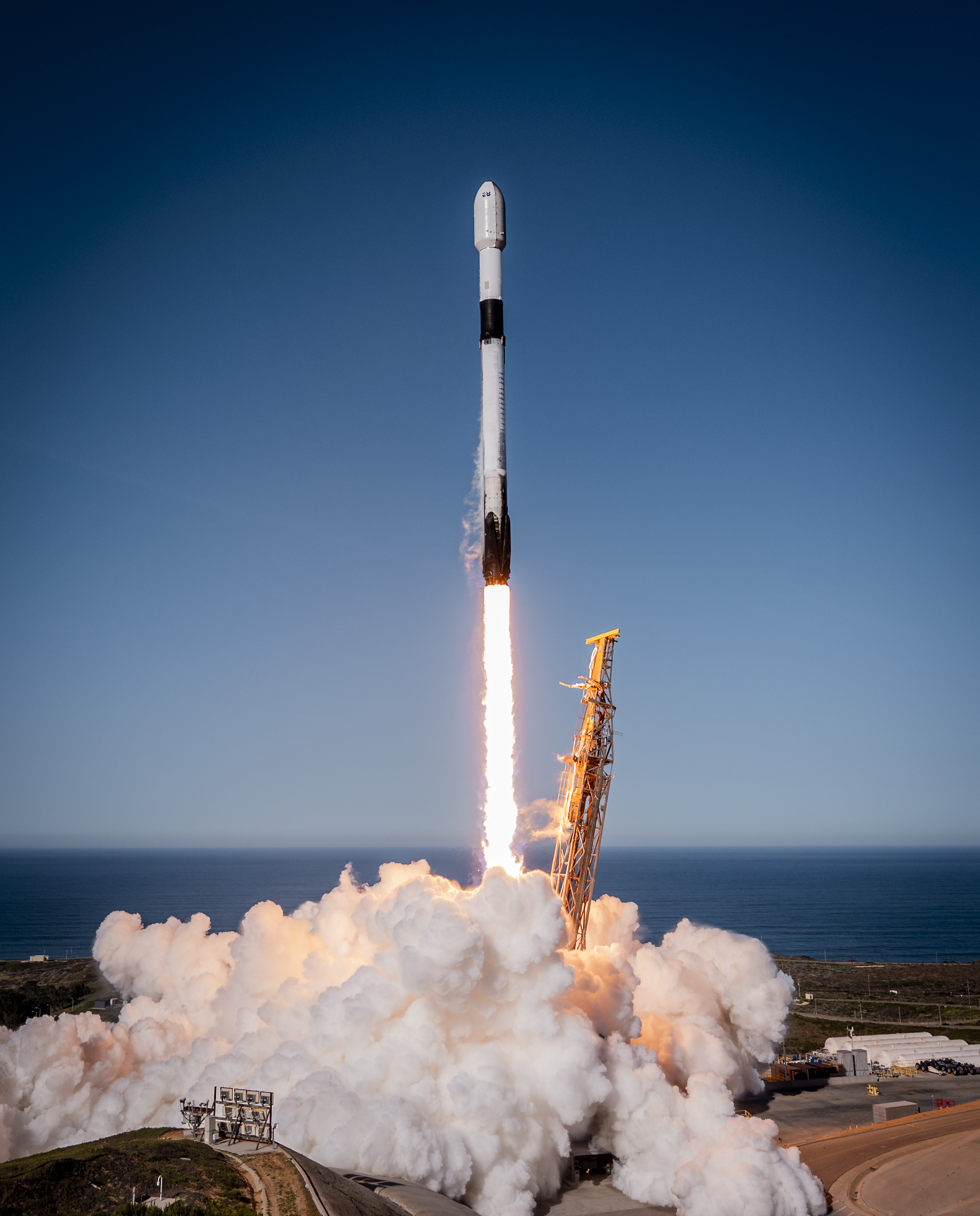 A white and black rocket launches into a clear blue sky backdropped by a body of water