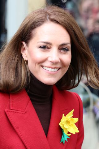 Kate Middleton wearing a red coat with a yellow daffodil pin smiling and looking at the camera