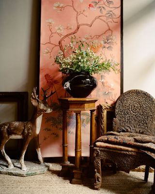 A glass vase in dark blue holds a bouquet of white flowers while sitting on a wooden side table positioned in front of a pink floral painted scene.