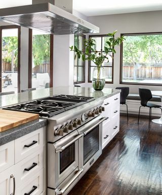 Large kitchen space with kitchen island and integrated stove, white cupboards, dark wood flooring, dining table in background with black dining chairs, white painted walls, dark wood framed windows