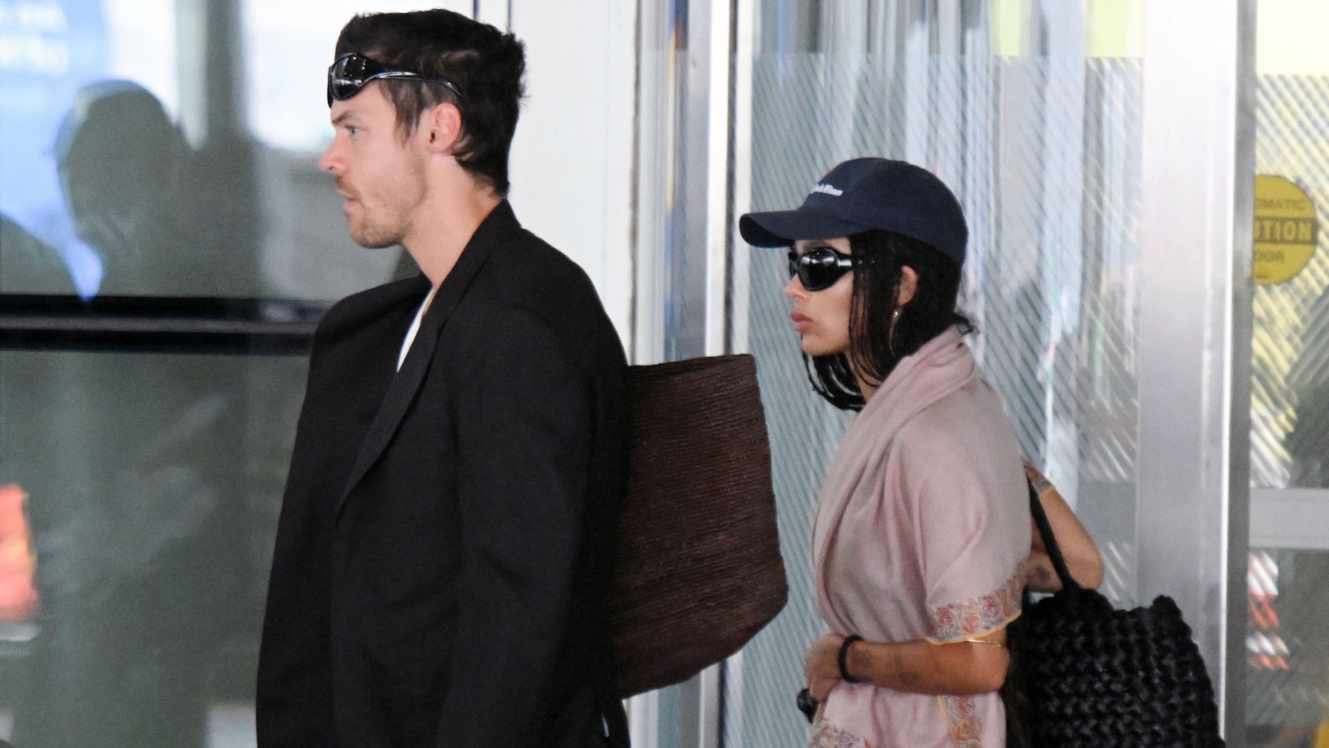 Zoe Kravitz and Harry Styles emerge from the airport. Zoe wears black trousers with heeled ballet pumps and a pink throw over her shoulders.