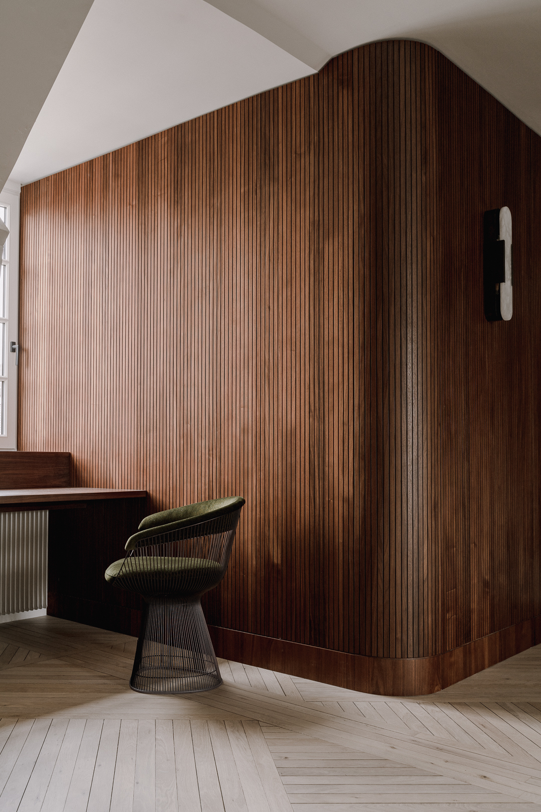 A refined attic apartment interior characterized by a warm, modernist aesthetic. The space is organized as an open-plan living area under a pitched roof, with exposed white-painted beams and triangular dormer windows that bring in soft natural light.