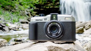 The Pentax 17 half-frame film camera in black and silver sitting on a rock with a waterfall behind it. 