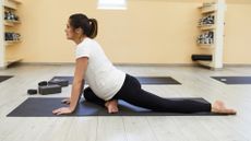 woman wearing white tshirt and black leggings performing a pigeon pose side on to the camera in a studio setting.