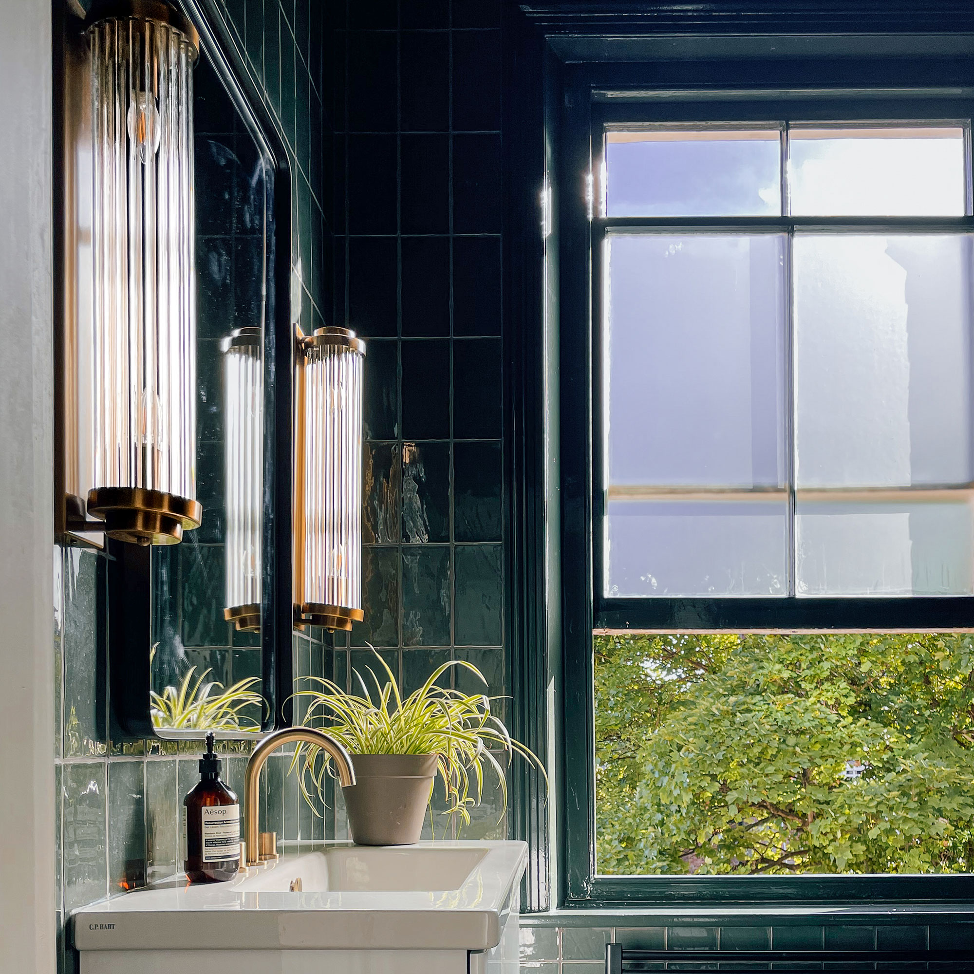 Dark green tiled bathroom with two glass and gold wall lights on either side of mirror above sink