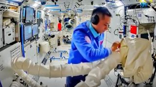 an astronaut in a blue flight suit examines cargo in a white bag aboard a white-walled space station