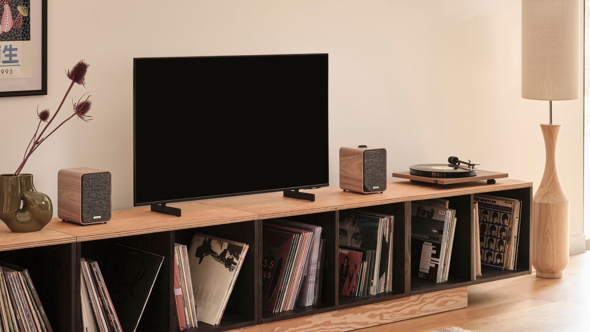 an image of a living room wall with an open fronted TV cabinet which has vinyls stored in it. On top of the cabinet is a TV and the Ruark speakers either side of it in walnut
