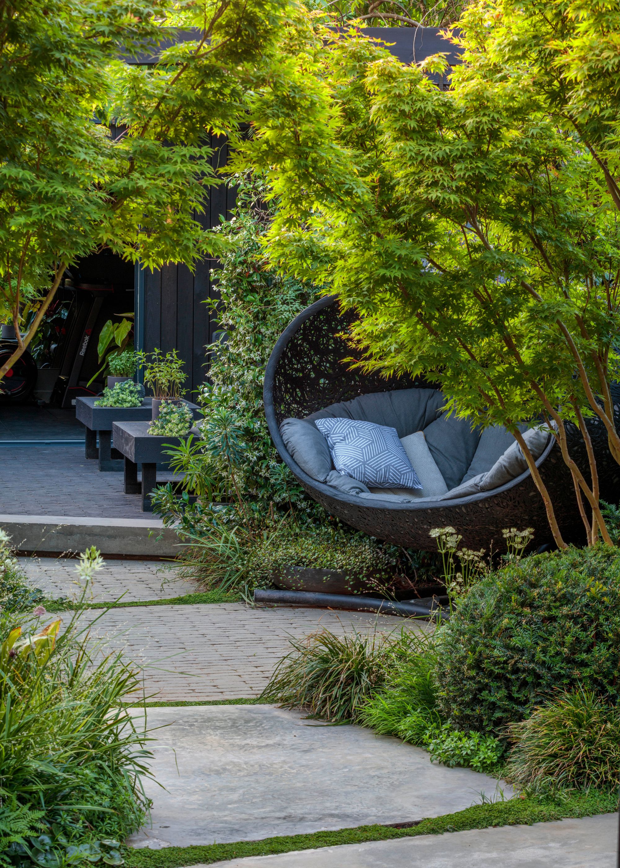 A garden with lots of lush greenery and paved ground. A black egg chair with gray cushions nestled into the greenery.