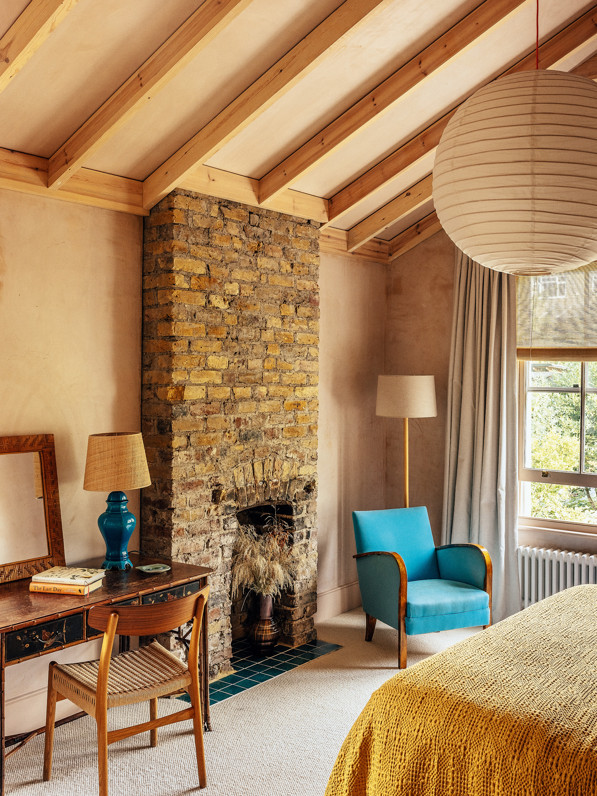 Bedroom with plaster pink walls, exposed rafters, exposed brick fireplace, vintage wood desk and blue armchair