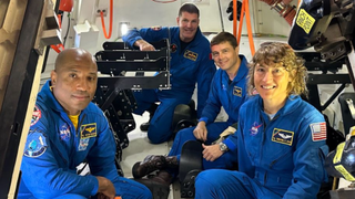 four astronauts in flight suits squatting inside of a spacecraft simulator surrounded by equipment