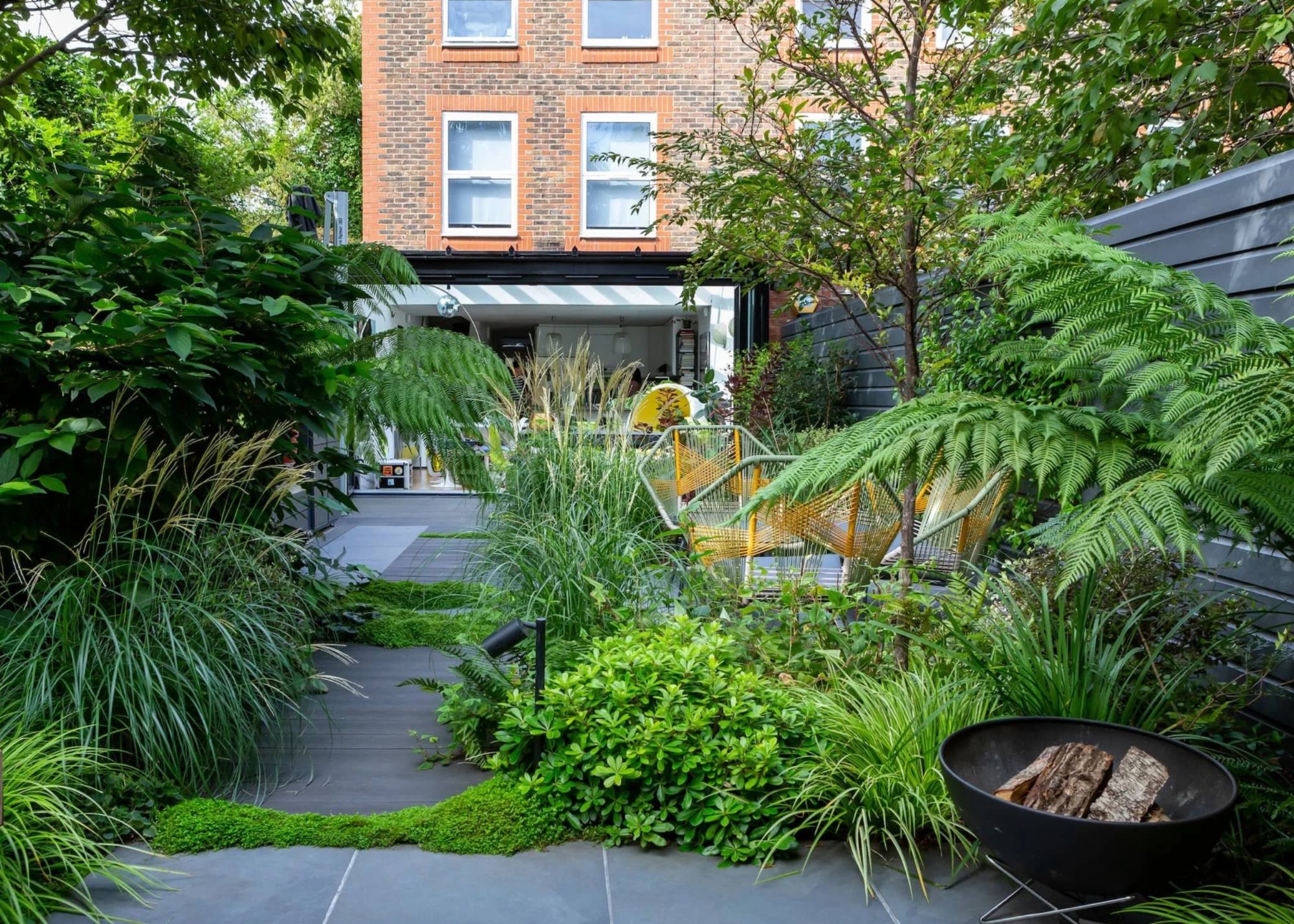 A calming modern urban garden with immersive repeated planting, zoned seating and a fire pit at the front